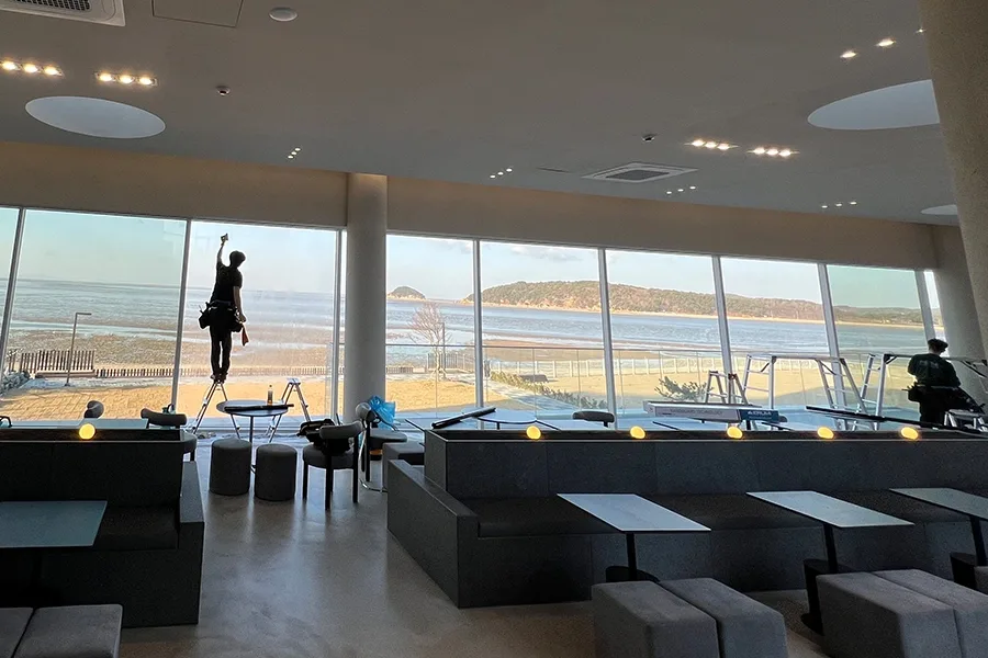 작업자가 사다리 위에서 해질녘 바다가 보이는 통유리에 필름을 부착하는 모습